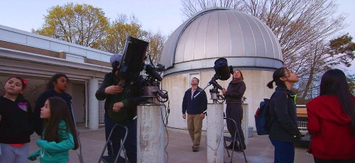 A group of children and adults observe the night sky using telescopes near an observatory dome.