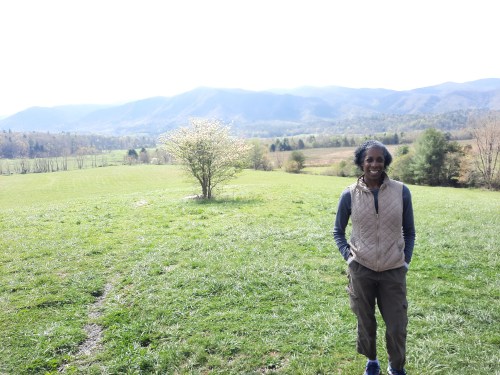 A person stands in a grassy field with mountains in the background and a tree nearby, enjoying a sunny day.