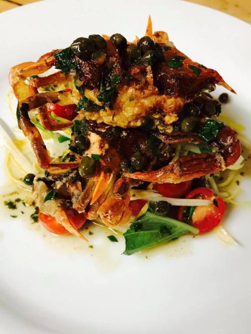 A plate of pasta topped with two crispy crabs, capers, and fresh herbs, garnished with cherry tomatoes and greens.
