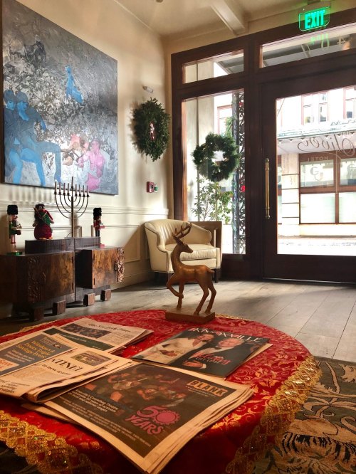 A cozy lobby with a festive table, newspapers, a reindeer statue, and holiday decorations, including a menorah and wreaths.