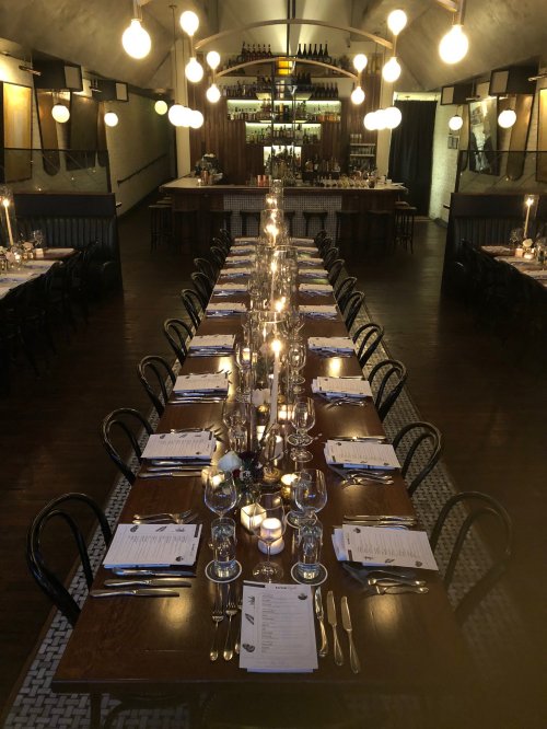 A long dining table set with candles and glassware in a warmly lit restaurant.