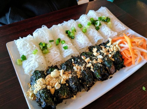 A plate of rice noodles topped with green onions, alongside grilled meat wrapped in leaves, garnished with peanuts and pickled vegetables.
