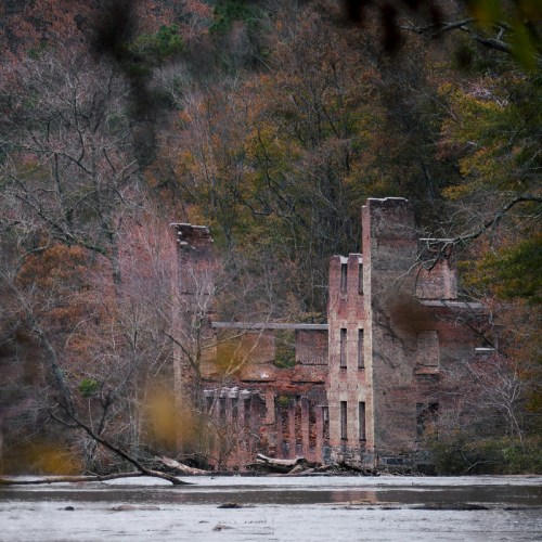 Ruins of a brick building surrounded by trees and a calm river, with autumn foliage in the background.