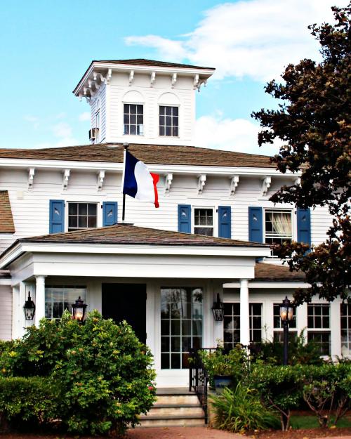 A charming white house with blue shutters and a French flag, surrounded by greenery under a blue sky.