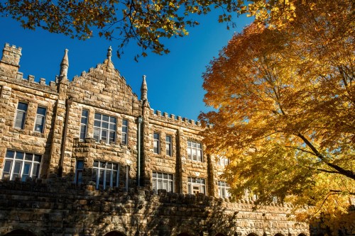 Historic stone building with gothic architecture, framed by vibrant autumn leaves against a clear blue sky.