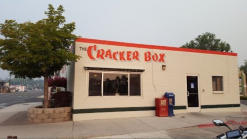 A small, white building with a red sign reading "The Cracker Box," featuring a tree and a newspaper stand outside.