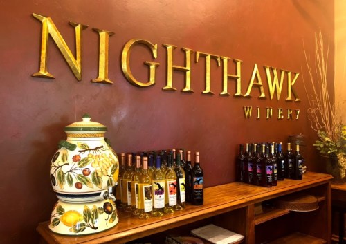 A wooden shelf displays various wine bottles beside a decorative ceramic container, with "Nighthawk Winery" sign above.