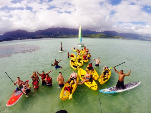 A group of people in yellow kayaks and paddleboards enjoying a sunny day on clear water with mountains in the background.