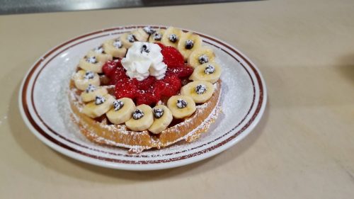 A delicious waffle topped with banana slices, strawberries, whipped cream, and chocolate chips, dusted with powdered sugar.