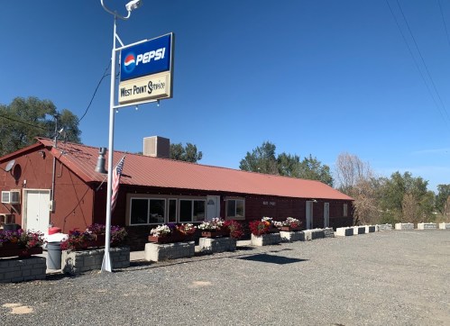 A red building with a Pepsi sign, surrounded by flowers and gravel, labeled "West Point Snyc." Clear blue sky above.