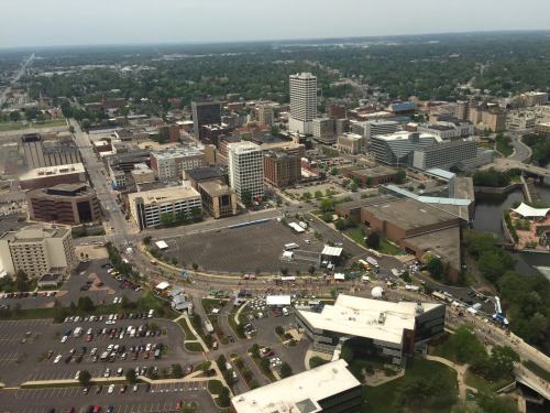 Aerial view of a cityscape featuring buildings, roads, and a river, with a large open area and parking lots visible.