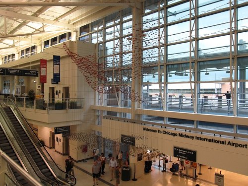 A spacious airport terminal with large windows, featuring a colorful art installation and travelers moving about.