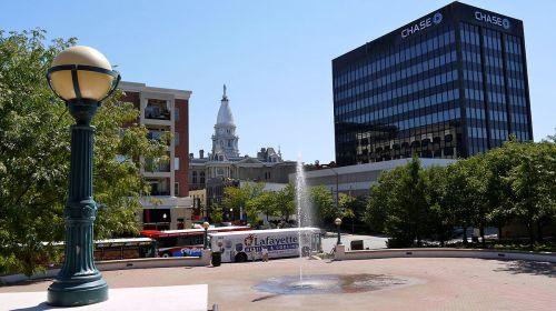 A city square featuring a fountain, trees, a bus, and buildings, including a Chase bank and a historic tower in the background.