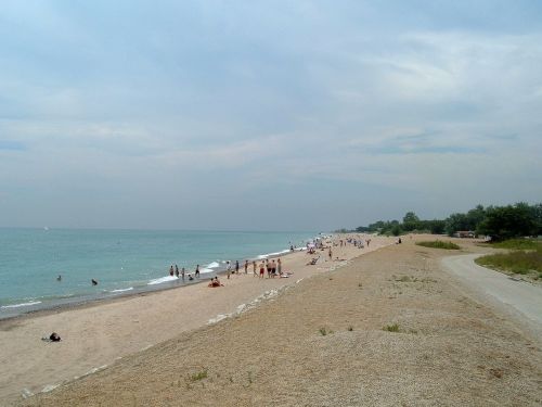 A sandy beach with people sunbathing and walking along the shore, under a cloudy sky.