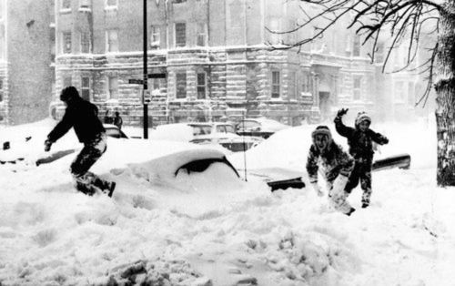 Children playing in deep snow, with a snow-covered car and buildings in the background during a winter storm.