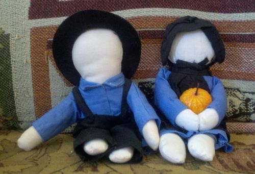 Two handmade dolls dressed in traditional Amish clothing, one holding a small orange pumpkin.