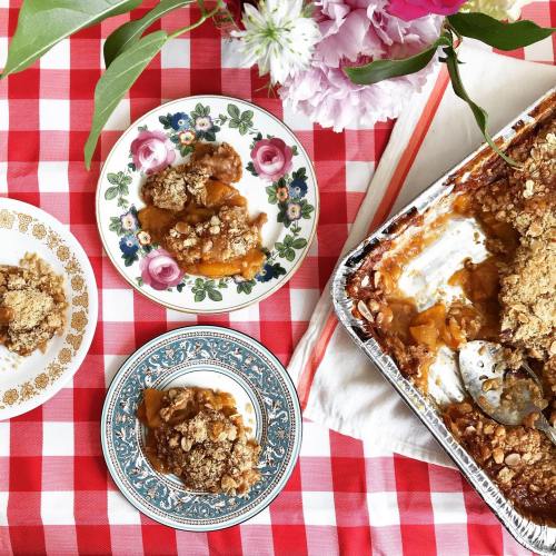 A table with a red checkered cloth featuring plates of peach cobbler and a baking dish, surrounded by flowers.