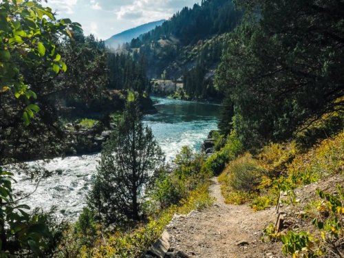 A scenic river view surrounded by lush greenery and mountains, with a winding path along the water's edge.