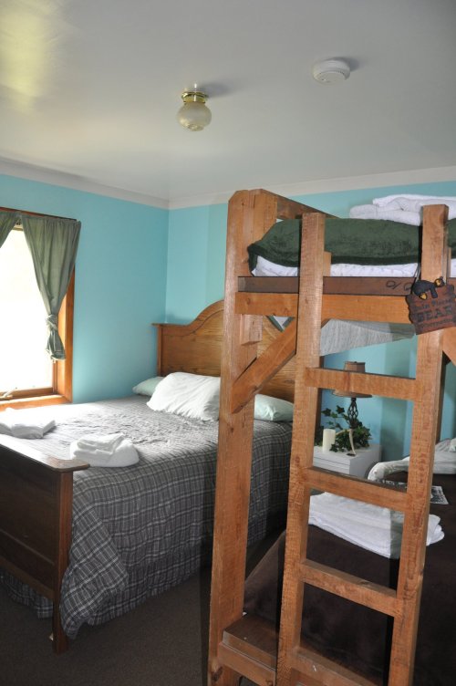 Cozy bedroom with a double bed and a wooden bunk bed, featuring light blue walls and natural light from a window.