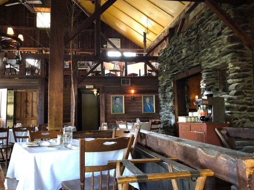 Cozy restaurant interior with wooden beams, stone walls, and tables set for dining, featuring two framed portraits.