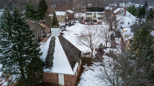 Aerial view of a snowy village with a church, houses, and trees, showcasing a peaceful winter landscape.