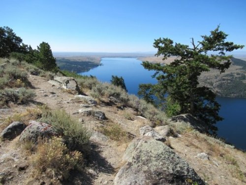 A scenic view of a lake surrounded by hills and trees, with a clear blue sky above.