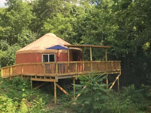 A cozy yurt on a wooden deck surrounded by lush greenery and trees. An umbrella provides shade on the deck.