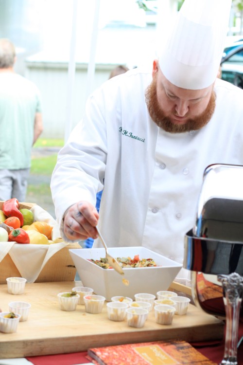 A chef in a white coat and hat prepares a dish, focusing on a bowl of food with small tasting cups nearby.