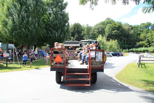 A trailer filled with people, including children, parked in a grassy area with trees and a crowd in the background.