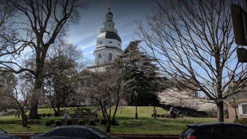 A historic building with a tall dome, surrounded by trees and blooming flowers, on a sunny day.