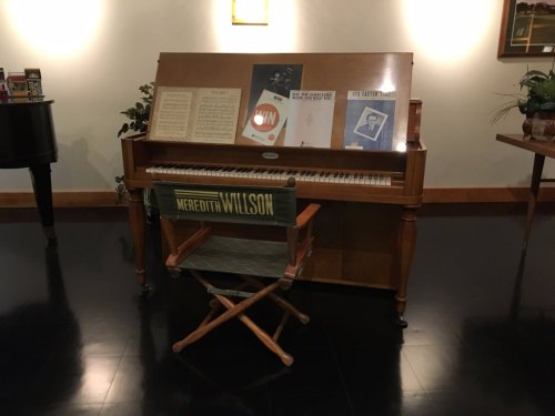 A wooden piano with sheet music and posters on top, accompanied by a director's chair labeled "Meredith Wilson."