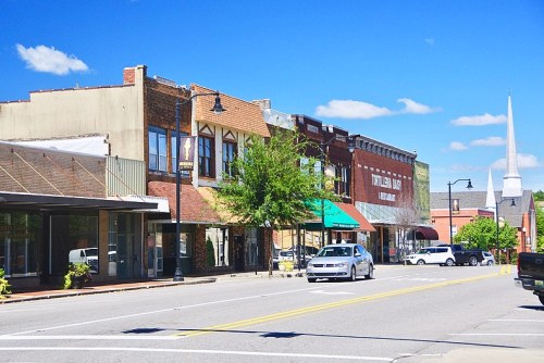 A sunny street view of a small town with historic buildings, shops, and a church steeple in the background.