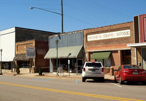A small town street featuring storefronts, including a music shop and a general store, under a clear blue sky.