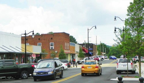 A street view of a small town with parked cars, shops, and streetlights under a cloudy sky.