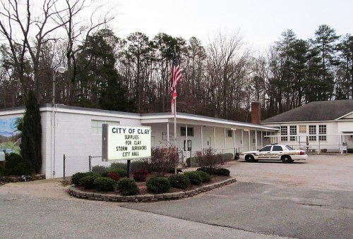 City Hall of Clay, featuring a sign for supplies and storm services, with a police car parked outside.