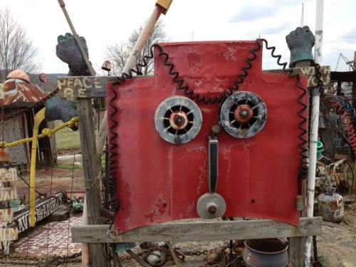 A whimsical sculpture made from metal parts, featuring a red face with circular eyes and a bell, surrounded by various objects.