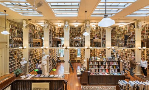 A spacious library with tall bookshelves, wooden floors, and large windows, featuring people browsing and statues among the books.