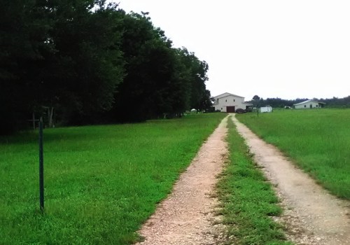 A gravel driveway leads through a grassy field to a house in the distance, surrounded by trees and open land.
