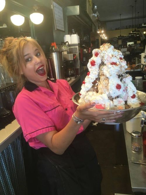 A smiling server in a pink shirt holds a large, extravagant ice cream sundae topped with whipped cream and cherries.
