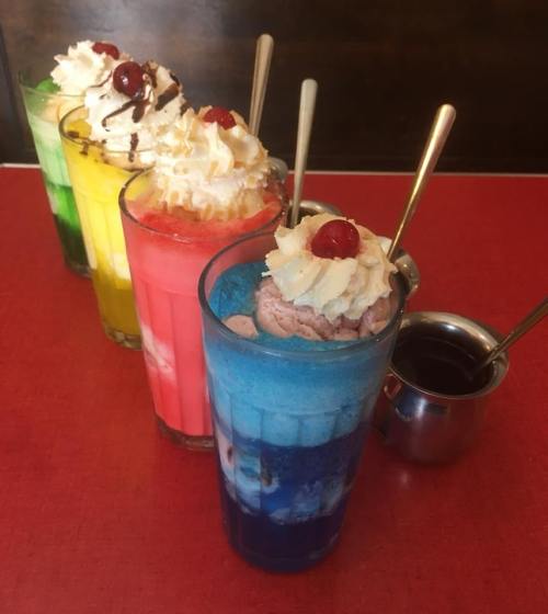 Four colorful ice cream sodas topped with whipped cream and cherries, served in tall glasses on a red table.