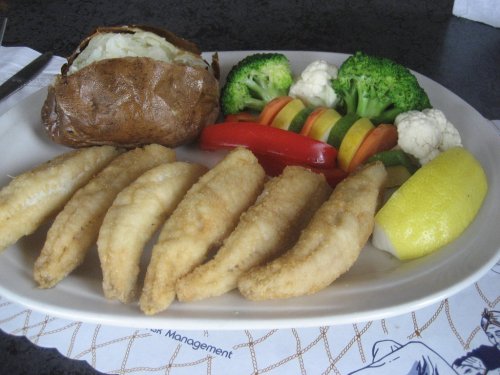A plate featuring fried fish fillets, a baked potato, and a colorful assortment of steamed vegetables.