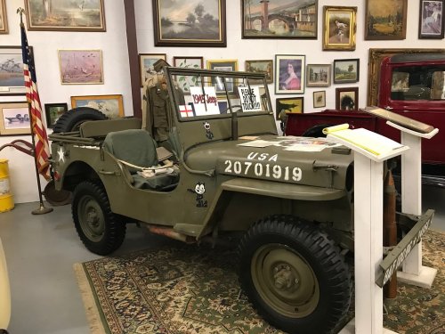 A vintage military jeep displayed in a museum, surrounded by paintings and artifacts, with an American flag in the background.