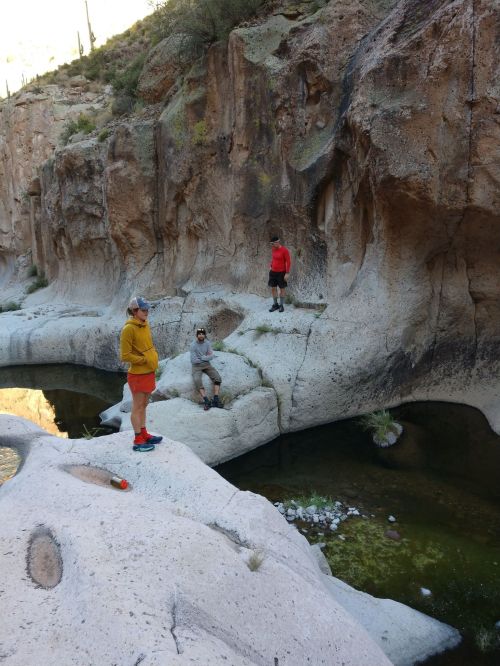 Three people stand on rocky terrain near a clear pool of water, surrounded by rugged cliffs and desert vegetation.