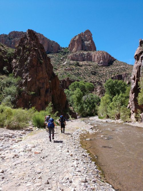 Hikers walking along a rocky riverbank with towering cliffs and greenery under a clear blue sky.