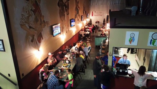 A lively restaurant scene with diners at tables, murals on the walls, and a bustling kitchen area in the background.