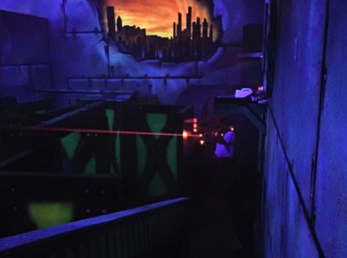 A dimly lit laser tag arena with neon green obstacles and a city skyline mural in the background.