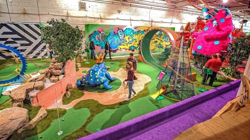 Colorful indoor mini-golf course featuring whimsical decorations, including a large dinosaur and vibrant murals.