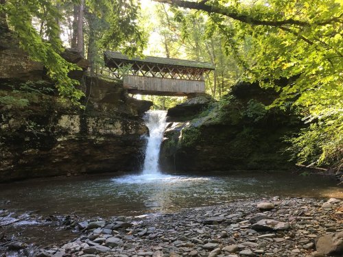 A serene waterfall flows beneath a wooden bridge, surrounded by lush greenery and rocky terrain.