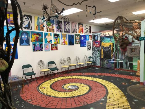 Colorful art-filled room with a spiral-patterned floor and chairs, featuring various paintings on the walls.