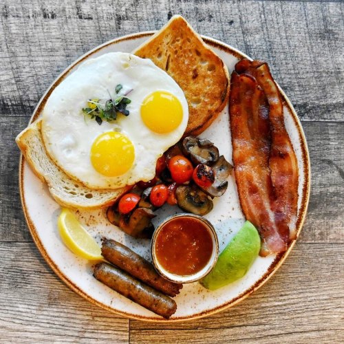 A plate of breakfast featuring two sunny-side-up eggs, toast, bacon, sausage, sautéed mushrooms, cherry tomatoes, and sauces.